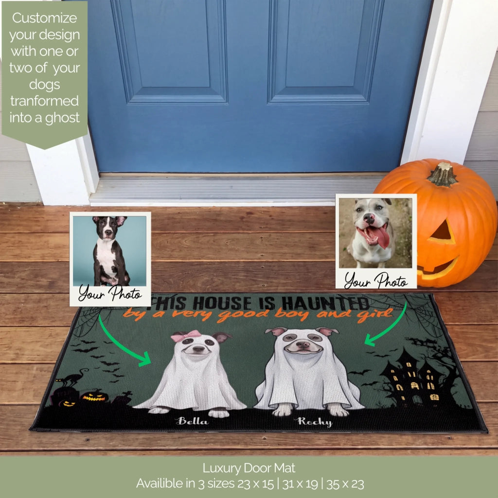 A personalized Halloween-themed luxury door mat displayed on a wooden porch in front of a blue door. The mat features cartoon illustrations of two dogs dressed as ghosts, with customizable text reading 'THIS HOUSE IS HAUNTED by a very good boy and girl'.