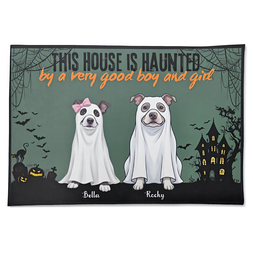 A rectangular Halloween-themed doormat with a dark green background, featuring the text 'THIS HOUSE IS HAUNTED by a very good boy and girl' above two dogs dressed as ghosts, with names 'Bella' and 'Recky' printed below them. The doormat is shown against a plain white background.