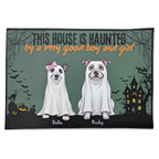 A rectangular Halloween-themed doormat with a dark green background, featuring the text 'THIS HOUSE IS HAUNTED by a very good boy and girl' above two dogs dressed as ghosts, with names 'Bella' and 'Recky' printed below them. The doormat is shown against a plain white background.