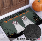 A Halloween-themed doormat featuring two dogs dressed as ghosts with the text 'THIS HOUSE IS HAUNTED by a very good boy and a girl'. The mat is shown on a tiled floor in front of a wooden door, with a pumpkin and a circular inset showing the mat's textured pattern.