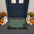 A Halloween-themed doormat displayed in front of a dark blue front door. The mat has a muted green background with black borders, a haunted house illustration, and the text 'THIS HOUSE IS HAUNTED by a very good ghost'.