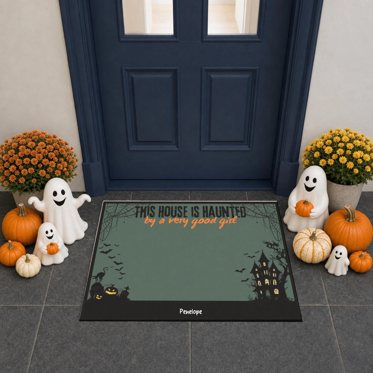 A Halloween-themed doormat displayed in front of a dark blue front door. The mat has a muted green background with black borders, a haunted house illustration, and the text 'THIS HOUSE IS HAUNTED by a very good ghost'.