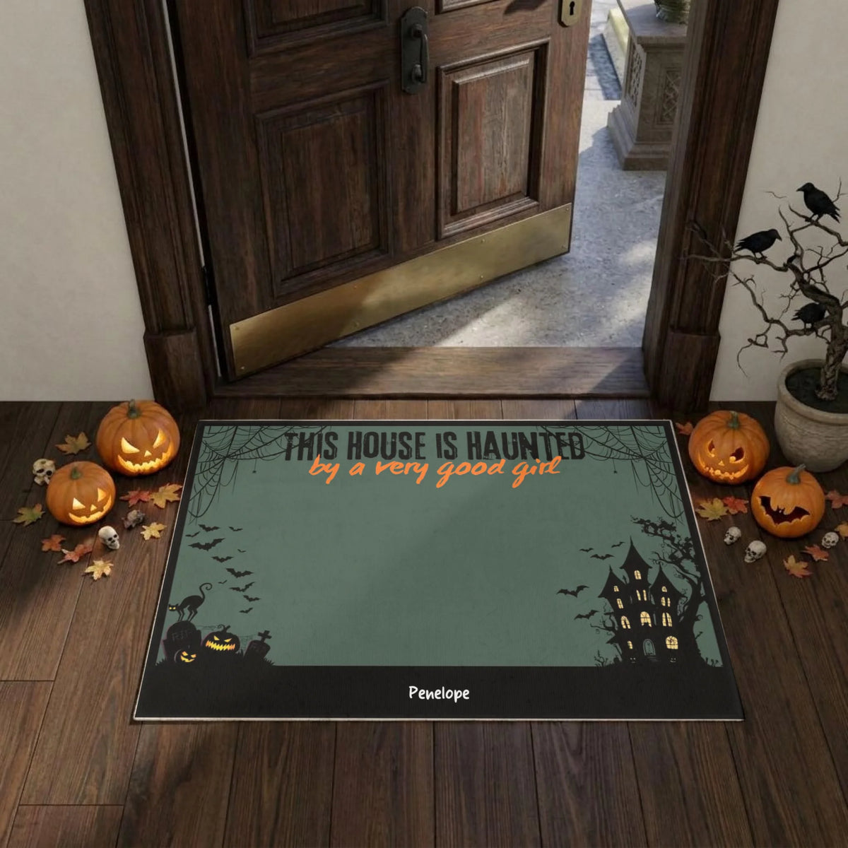 A Halloween-themed doormat placed in front of an open wooden door on a wood floor, surrounded by pumpkins, leaves, and small skulls. The mat has a muted green background with black silhouettes of a haunted house, bats, and a cat, plus orange text reading 'THIS HOUSE IS HAUNTED by a very good ghost.