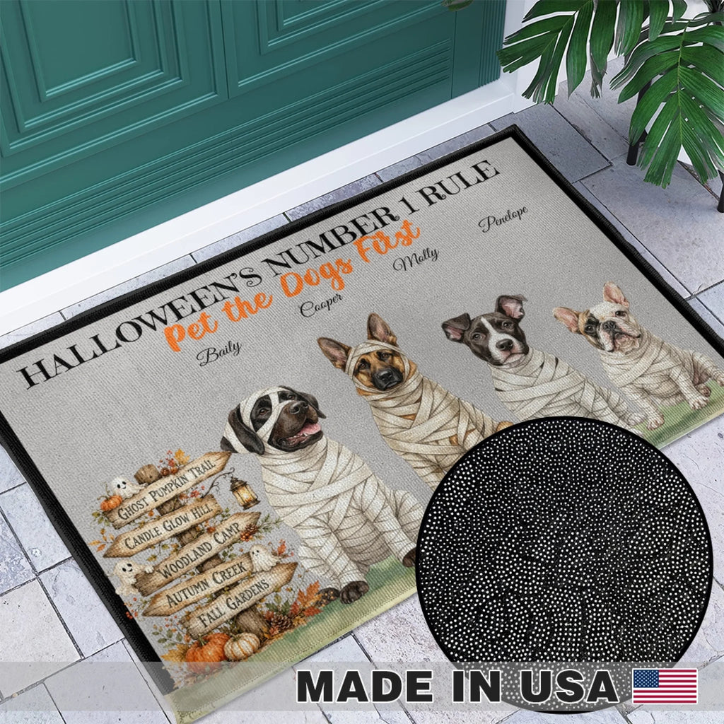 A rectangular doormat with a Halloween-themed design featuring four mummy dogs, the text 'HALLOWEEN'S NUMBER 1 RULE' and 'Pet the Dogs First', plus decorative fall signs and pumpkins. The mat is shown placed at a doorway on a tiled floor, with a black circular inset highlighting the printed pattern detail and a 'MADE IN USA' badge at the bottom.
