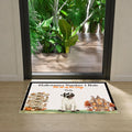 A rectangular Halloween-themed doormat placed in front of a glass door. The mat has a white background with a black border, a cartoon pug dressed as a mummy, and seasonal illustrations including a haunted house, pumpkins, autumn leaves, and directional signs.