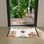 A rectangular Halloween-themed doormat placed in front of a glass door. The mat has a white background with a black border, a cartoon pug dressed as a mummy, and seasonal illustrations including a haunted house, pumpkins, autumn leaves, and directional signs.