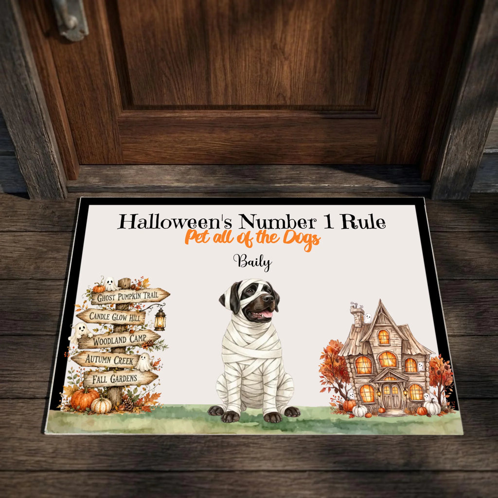 A personalized Halloween doormat placed in front of a dark wooden door. The mat features a white background with a black border, an illustration of a dog wrapped in bandages, a haunted house, autumn leaves, pumpkins, and the text 'Halloween's Number 1 Rule' and the name 'Baily'.