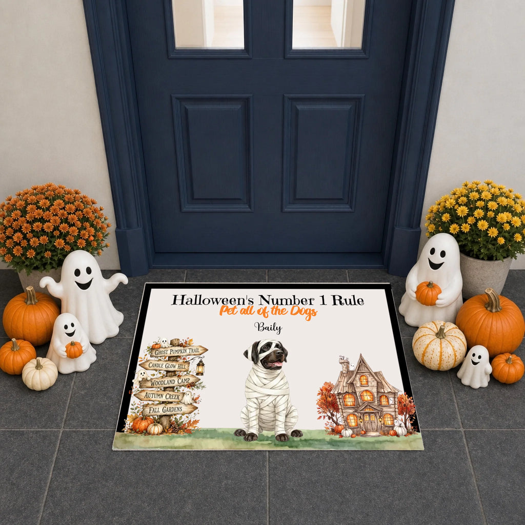 A rectangular Halloween-themed doormat displayed in front of a dark blue front door, surrounded by fall decorations including pumpkins, ghost figurines, and mums. The mat features a dog dressed as a mummy, a haunted house, a directional signpost, and the text 'Halloween's Number 1 Rule' and 'Pet all of the Dogs - Daily'.