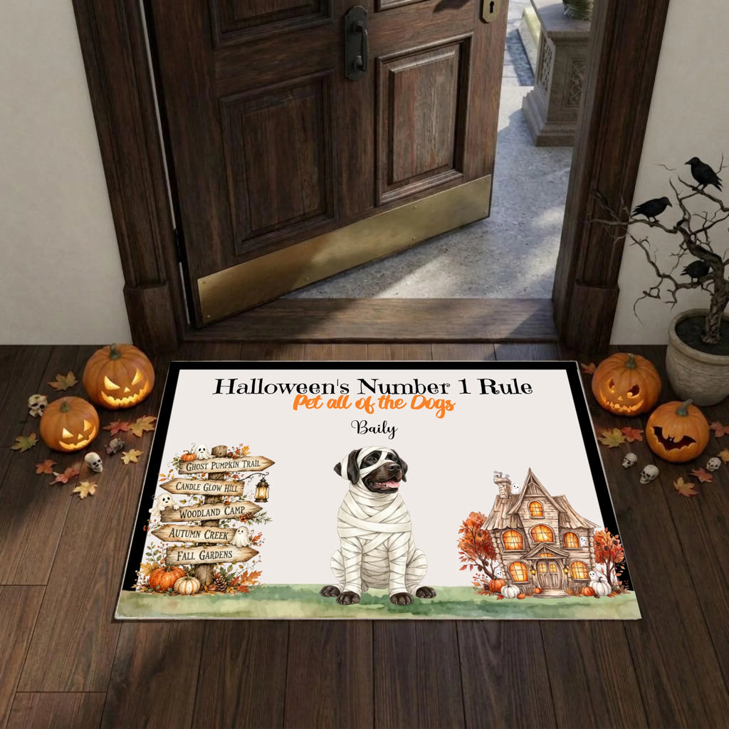 A personalized Halloween-themed doormat displayed on a wooden entryway floor. The mat features a custom design with the text 'Halloween's Number 1 Rule' and 'Pet all of the Dogs - Bailey', accompanied by an illustration of a mummy dog, a haunted house, and a directional signpost.