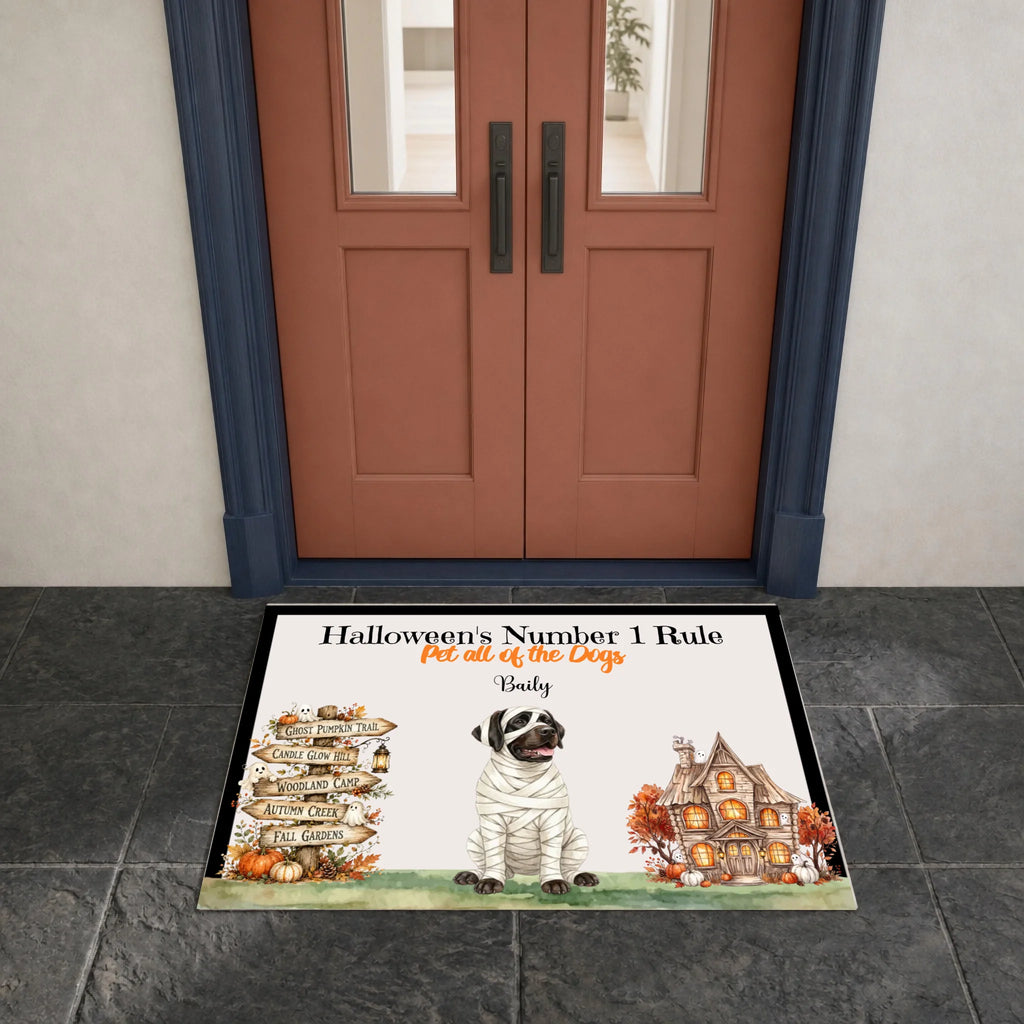 A personalized Halloween doormat displayed in front of a double door. The doormat features a white background with a black border, a cartoon dog dressed as a bandaged zombie, a stack of autumn leaves pointing in different directions, and a haunted house.