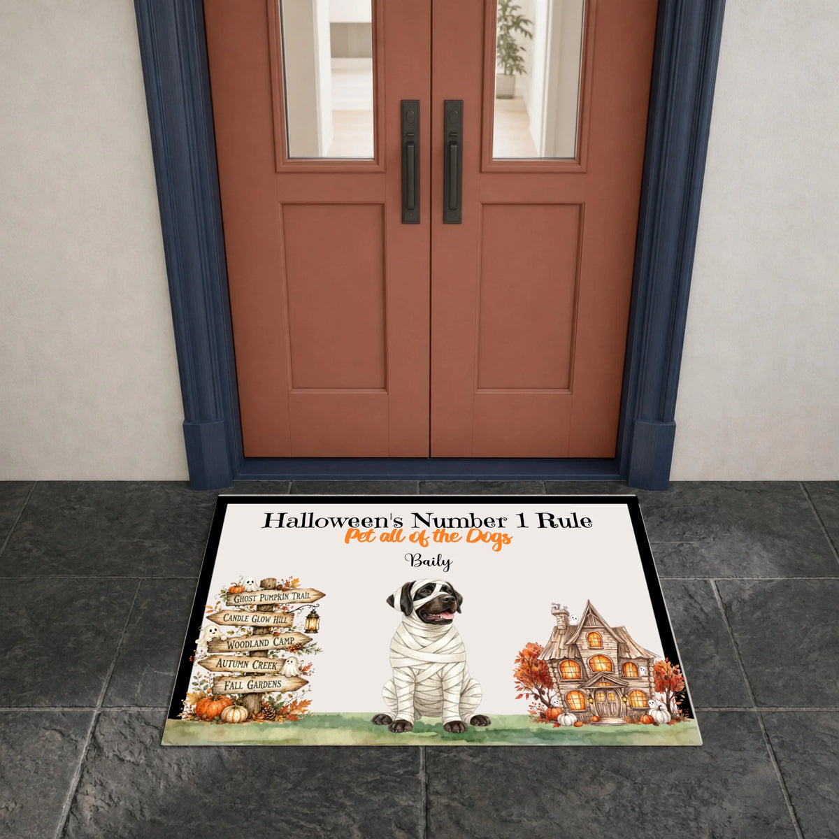 A personalized Halloween doormat displayed in front of a double door. The doormat features a white background with a black border, a cartoon dog dressed as a bandaged zombie, a stack of autumn leaves pointing in different directions, and a haunted house.
