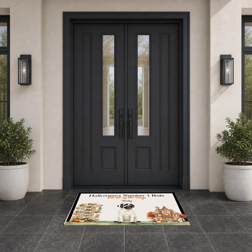 A rectangular front door doormat with a Halloween-themed illustration and the text 'Halloween Number 1 Bole' and 'Stay in the Fun' is placed in front of dark charcoal double front doors on a tiled entryway floor, flanked by potted plants and wall sconces.