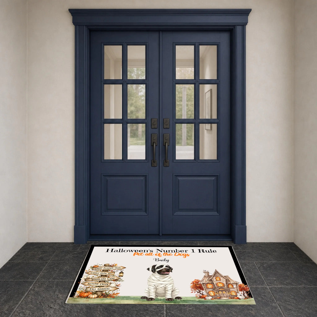 A Halloween-themed doormat with the text 'Halloween's Number 1 Rule' and a cartoon dog dressed as a mummy, placed in front of dark blue double doors on a gray tiled floor.