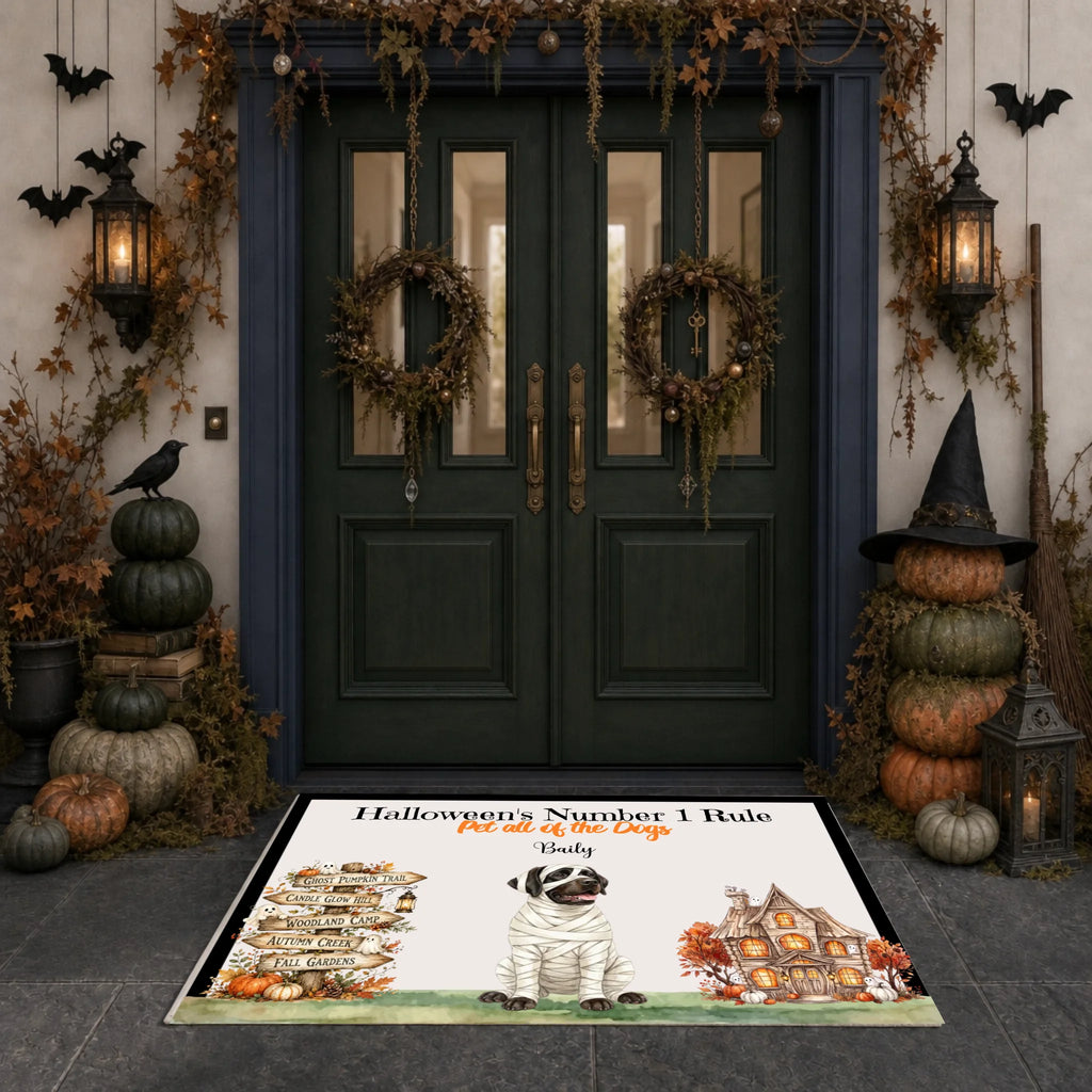 A rectangular Halloween-themed doormat displayed in front of a dark green double door decorated with wreaths, pumpkins, lanterns, and bats. The mat features a mummy dog illustration and the text 'Halloween's Number 1 Rule' and 'Beasty'.