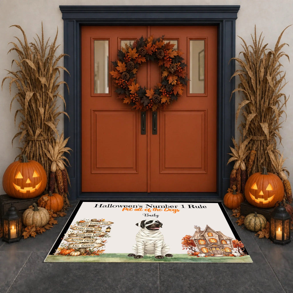 A Halloween-themed doormat displayed on a dark stone entryway in front of a rustic double door decorated with a wreath, pumpkins, corn, and lanterns. The mat features a mummy dog, a haunted house, and the text 'Halloween's Number 1 Rule Put all of the cats out of the house' with the name 'Emily' written below.