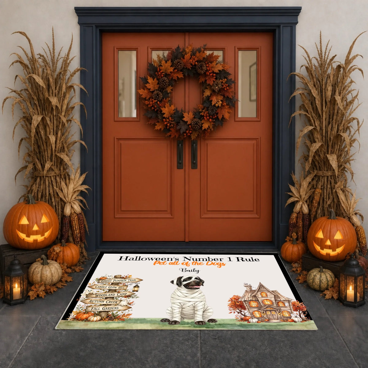 A Halloween-themed doormat displayed on a dark stone entryway in front of a rustic double door decorated with a wreath, pumpkins, corn, and lanterns. The mat features a mummy dog, a haunted house, and the text 'Halloween's Number 1 Rule Put all of the cats out of the house' with the name 'Emily' written below.