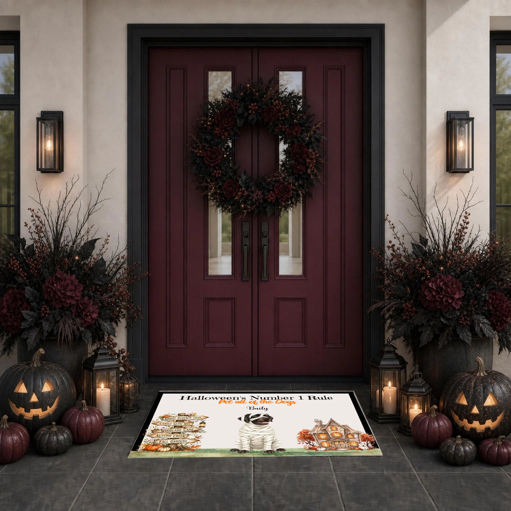 A Halloween-themed doormat displayed on a dark tiled entryway floor in front of a burgundy double door with a wreath. The mat features a cartoon bat, haunted houses, pumpkins, and the text 'Halloween Nightmare Mode' and 'Stay Up Late'.