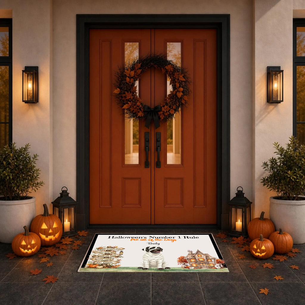 A Halloween-themed doormat with a haunted house illustration and the text 'Halloween Number 1 Rule: Stay Home' is displayed on a dark tiled entryway floor in front of double doors decorated with a wreath, pumpkins, and lanterns.