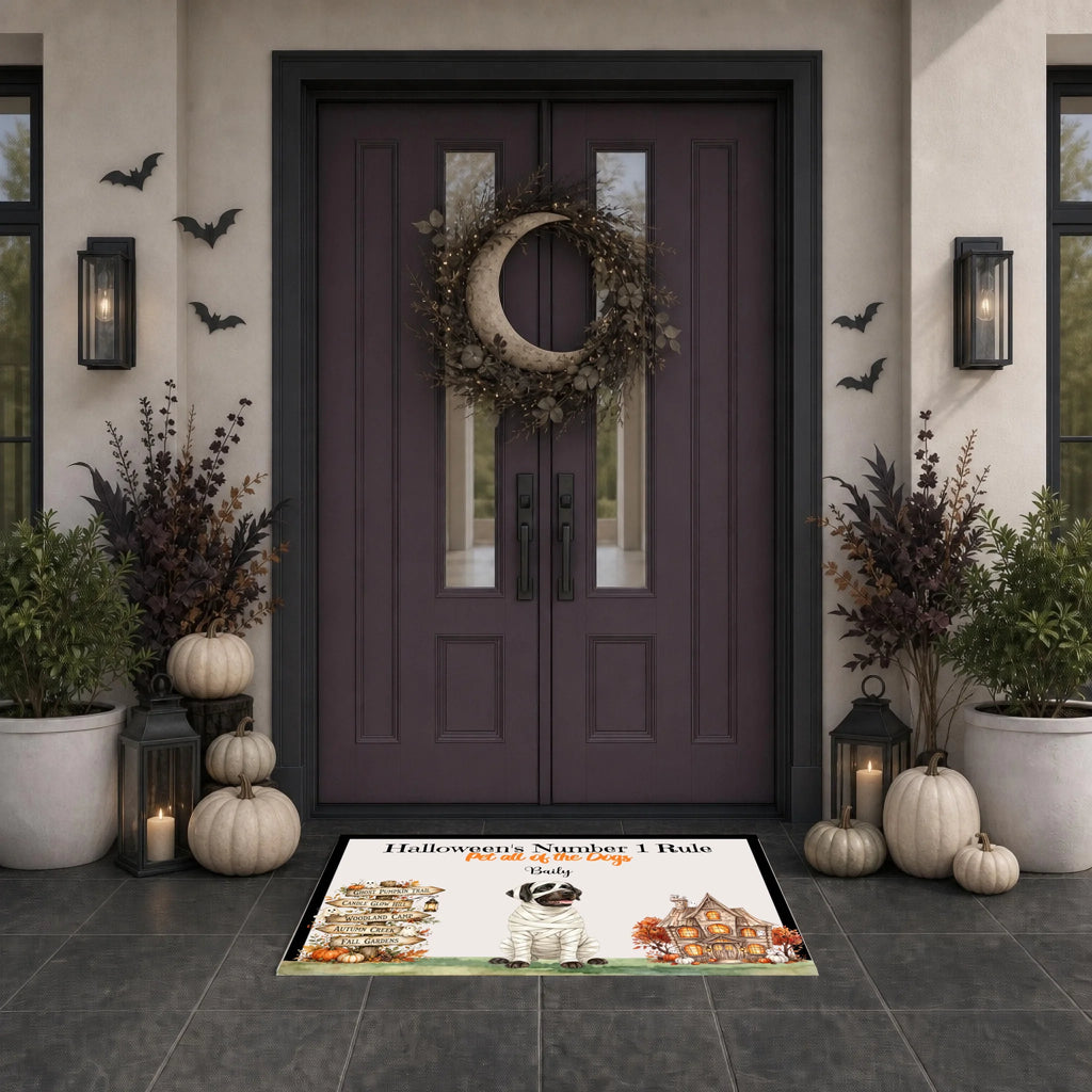 A Halloween-themed doormat with illustrations of a haunted house, a skull, and the text 'Halloween Number 1 Rule: Stay Home' is displayed on a dark tiled entryway floor in front of a dark purple double door decorated with a wreath, pumpkins, and lanterns.