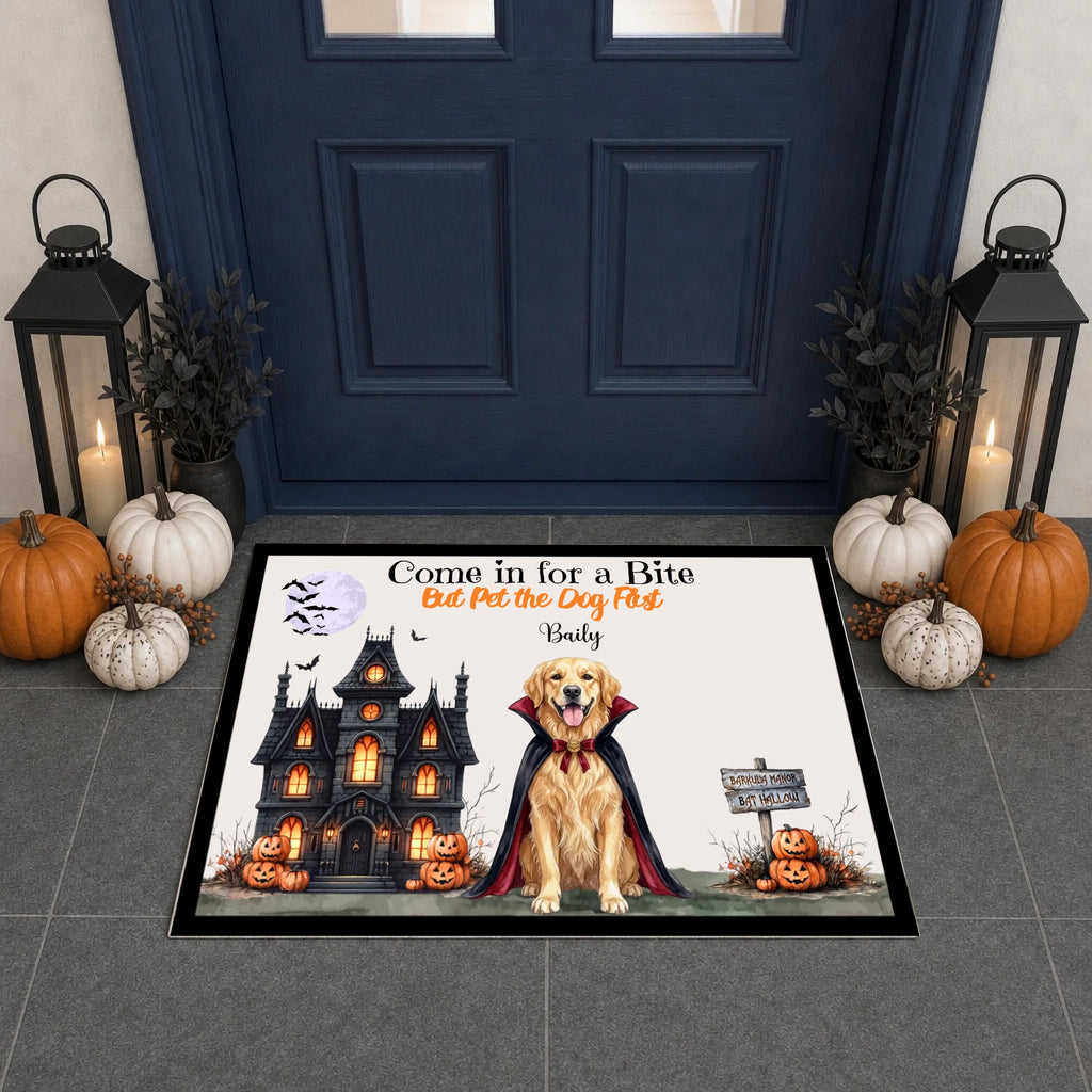 A rectangular Halloween-themed doormat displayed in front of a dark blue front door. The mat features a golden retriever dressed as a vampire in front of a haunted house, with pumpkins, bats, and the text 'Come in for a Bite Eat Pet the Dog First: Baby'.