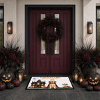 A Halloween-themed doormat featuring a vampire cat and haunted house illustration, placed in front of a dark red front door decorated with a wreath, pumpkins, lanterns, and floral arrangements.