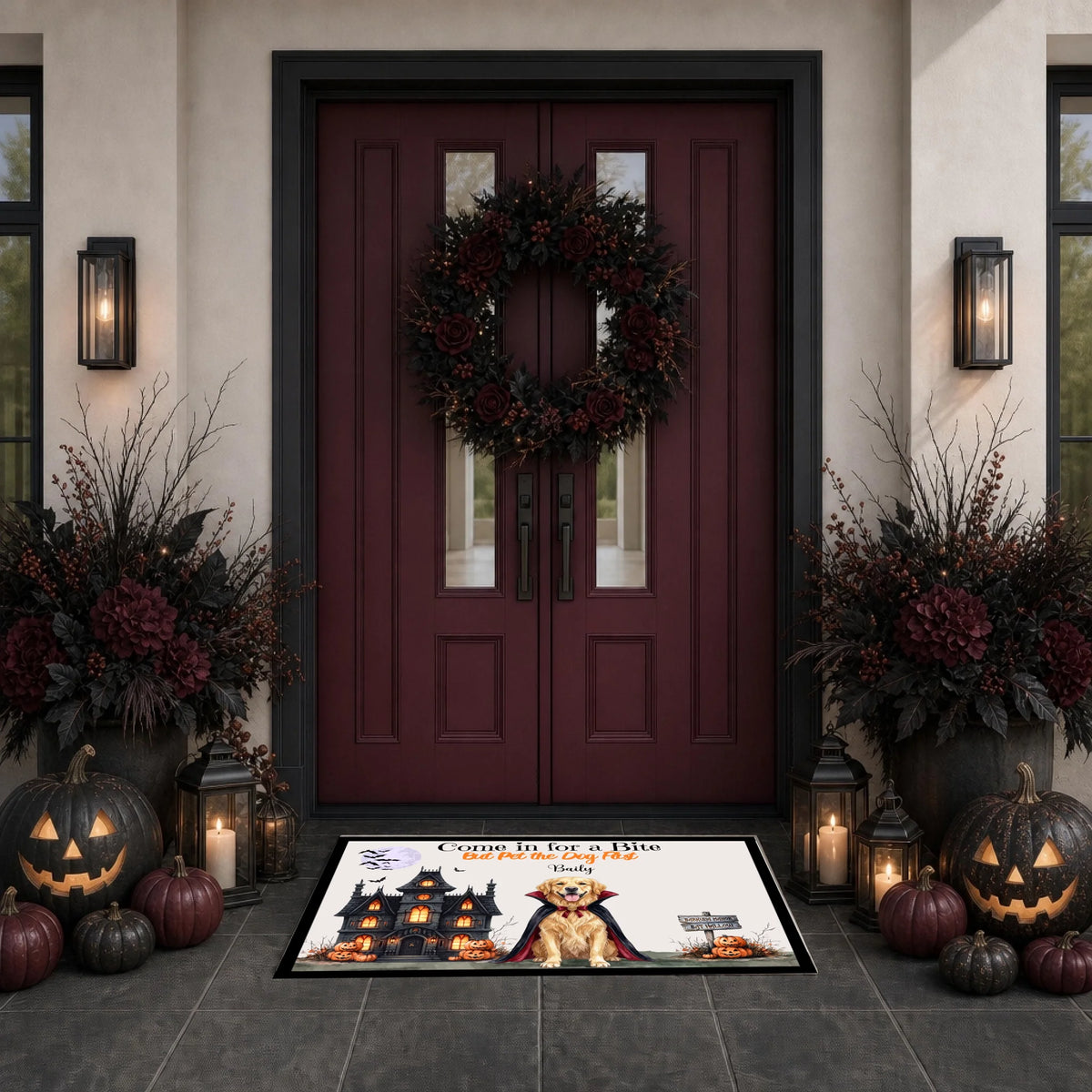 A Halloween-themed doormat featuring a vampire cat and haunted house illustration, placed in front of a dark red front door decorated with a wreath, pumpkins, lanterns, and floral arrangements.