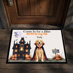 A rectangular Halloween-themed doormat placed in front of a dark wood door. The mat features a golden retriever dressed as a vampire, a haunted house, pumpkins, bats, and the text 'Come in for a Bite But Pet the Dog First Badly'.