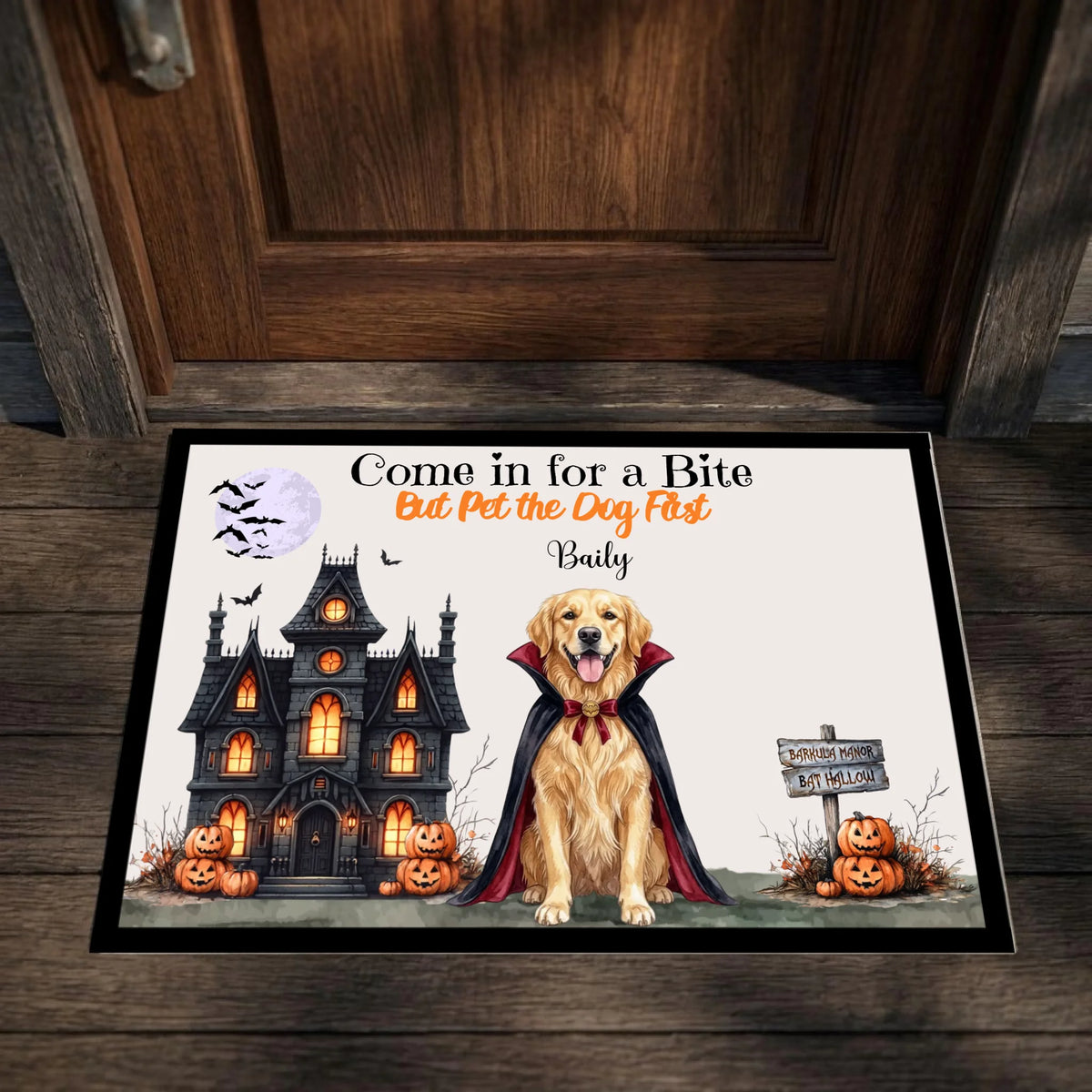 A rectangular Halloween-themed doormat placed in front of a dark wood door. The mat features a golden retriever dressed as a vampire, a haunted house, pumpkins, bats, and the text 'Come in for a Bite But Pet the Dog First Badly'.