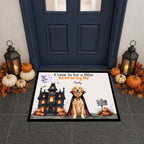 A rectangular Halloween-themed doormat displayed on a tiled entryway floor in front of a dark blue door. The mat features an illustration of a golden retriever dressed as a vampire in front of a haunted house, with pumpkins, bats, and the text 'Come in for a Bite But Pet the Dog First' and 'Buddy'.