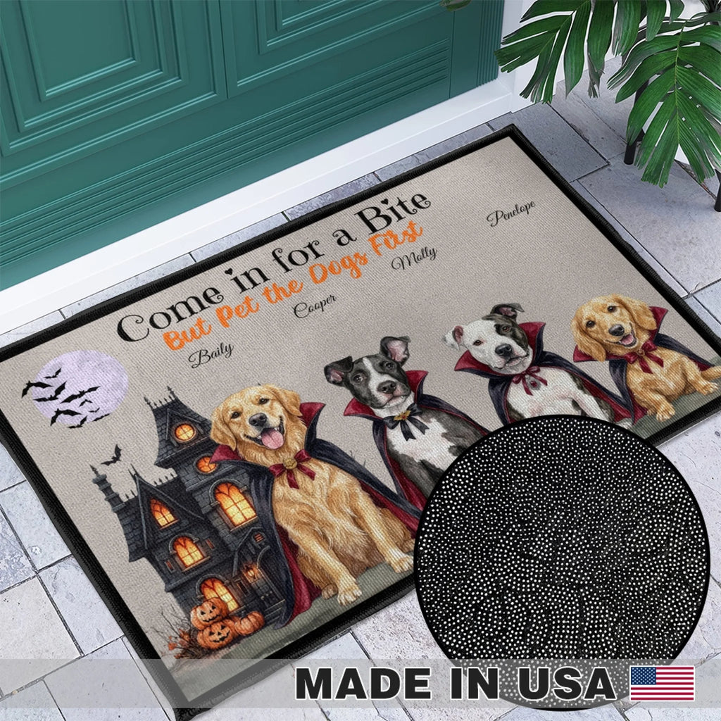 Personalized Halloween doormat shown in a front porch setting with a teal door and tiled floor. The mat features a haunted house, pumpkins, vampire dogs, and the printed phrase “Come in for a Bite But Pet the Dogs First,” plus customizable dog names.