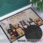 Personalized Halloween doormat shown in a front porch setting with a teal door and tiled floor. The mat features a haunted house, pumpkins, vampire dogs, and the printed phrase “Come in for a Bite But Pet the Dogs First,” plus customizable dog names.