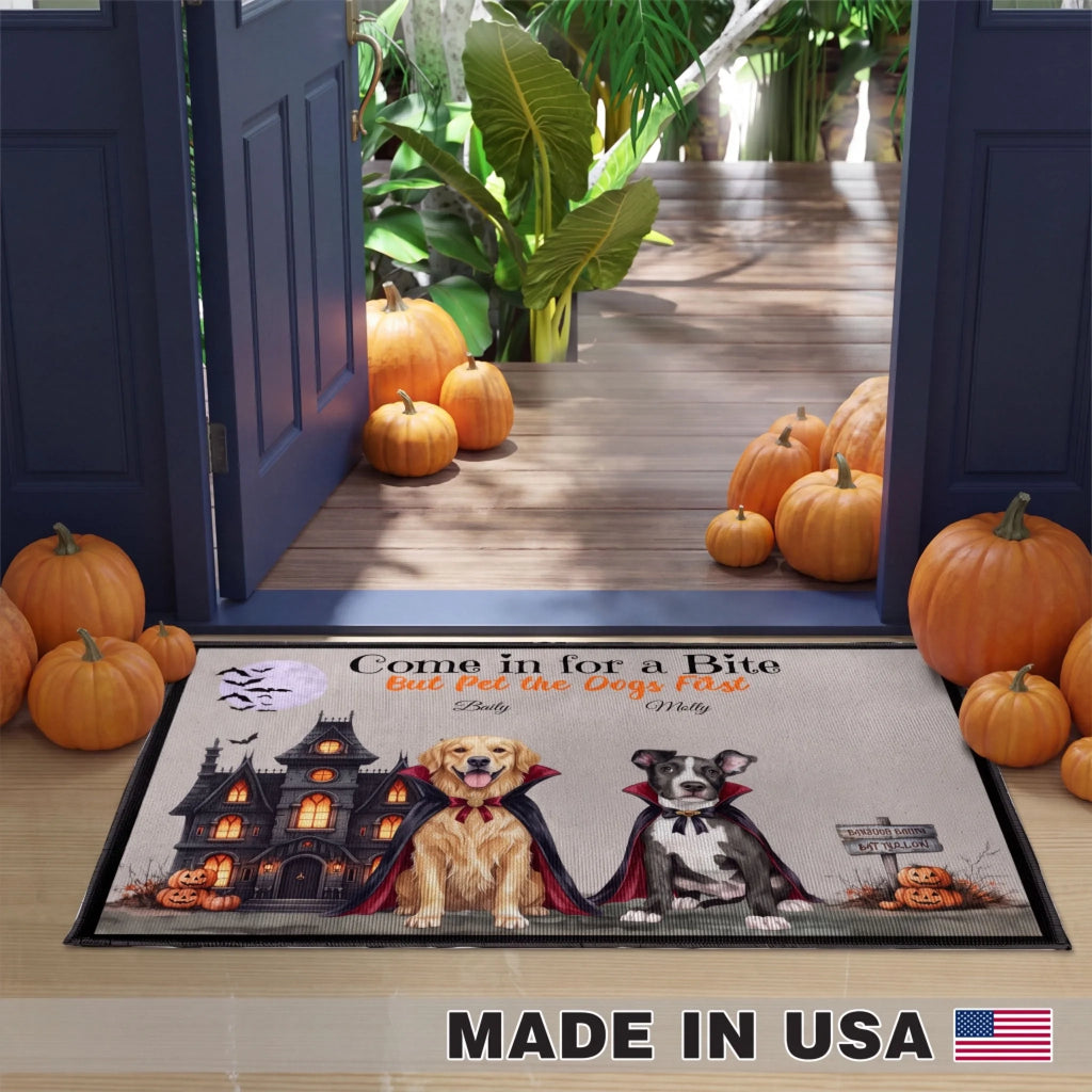 A rectangular doormat with a Halloween-themed illustration of two vampire dogs and a haunted house, placed in front of open dark blue doors on a porch with pumpkins around it. The mat includes the text 'Come in for a Bite But Pet the Dogs First' and a 'MADE IN USA' banner appears at the bottom of the image.