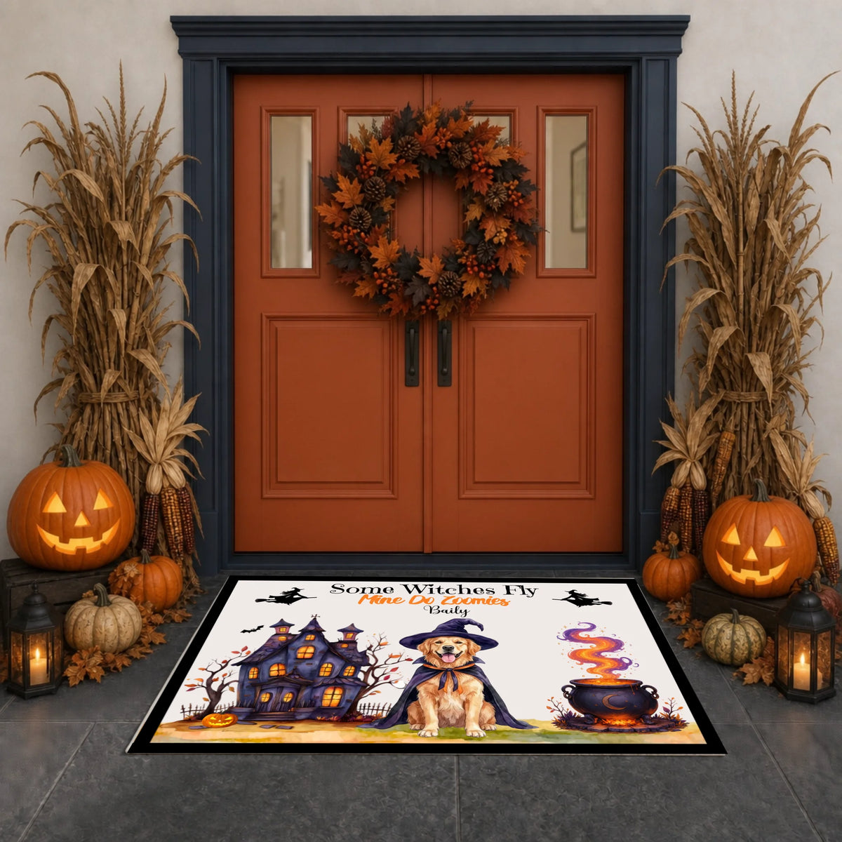 A Halloween-themed doormat featuring a witch cat, haunted house, cauldron, and bats, placed in front of a dark blue double door decorated with a wreath, pumpkins, cornstalks, and lanterns.