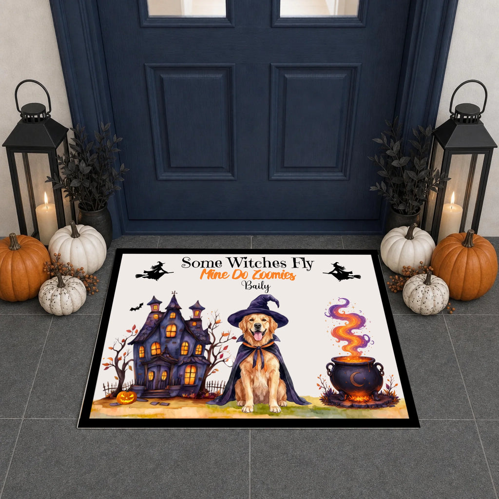A rectangular Halloween-themed doormat displayed in front of a dark blue front door. The mat features a golden retriever dog in a witch hat and cape next to a haunted house and a cauldron, with the text 'Some Witches Fly Mine Or Yours Daily' and the name 'Emily' printed on it.