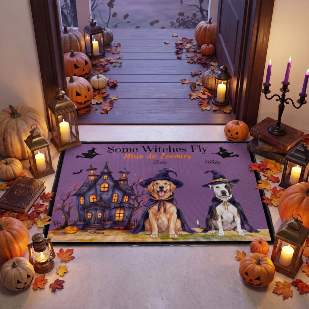 A Halloween-themed rectangular floor mat displayed on a porch threshold, surrounded by pumpkins, lanterns, and autumn leaves. The mat features a purple sky with the text 'Some Witches Fly Mine do Scoops' and illustrations of two dogs in witch hats flying in front of a haunted house.