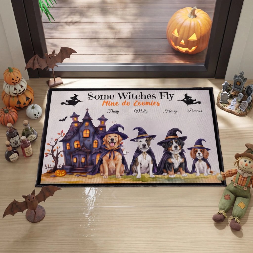 A rectangular Halloween-themed doormat displayed on a light wood porch floor in front of a window. The mat features a haunted house, four dogs wearing witch hats, bats, and the text 'Some Witches Fly Mine do Eomies' with placeholder names below.