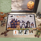 A Halloween-themed luxury doormat displayed on a wooden porch floor, surrounded by seasonal props like a carved pumpkin, bottles, a scarecrow, and a bat figurine. The mat features four dogs dressed as witches in front of a haunted house, with customizable text and four sample dog photo inserts shown below the main mat.