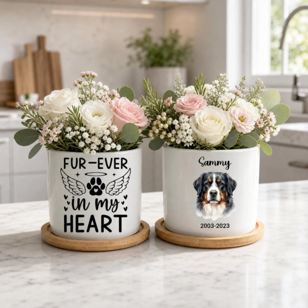Two white ceramic flower pots with wooden bases sit on a marble countertop in a bright kitchen. Each pot holds a floral arrangement and is printed with memorial-style pet text: the left pot reads “FUR-EVER in my HEART” with a paw print and wings, and the right pot reads “Sammy 2003-2023” with a dog portrait.