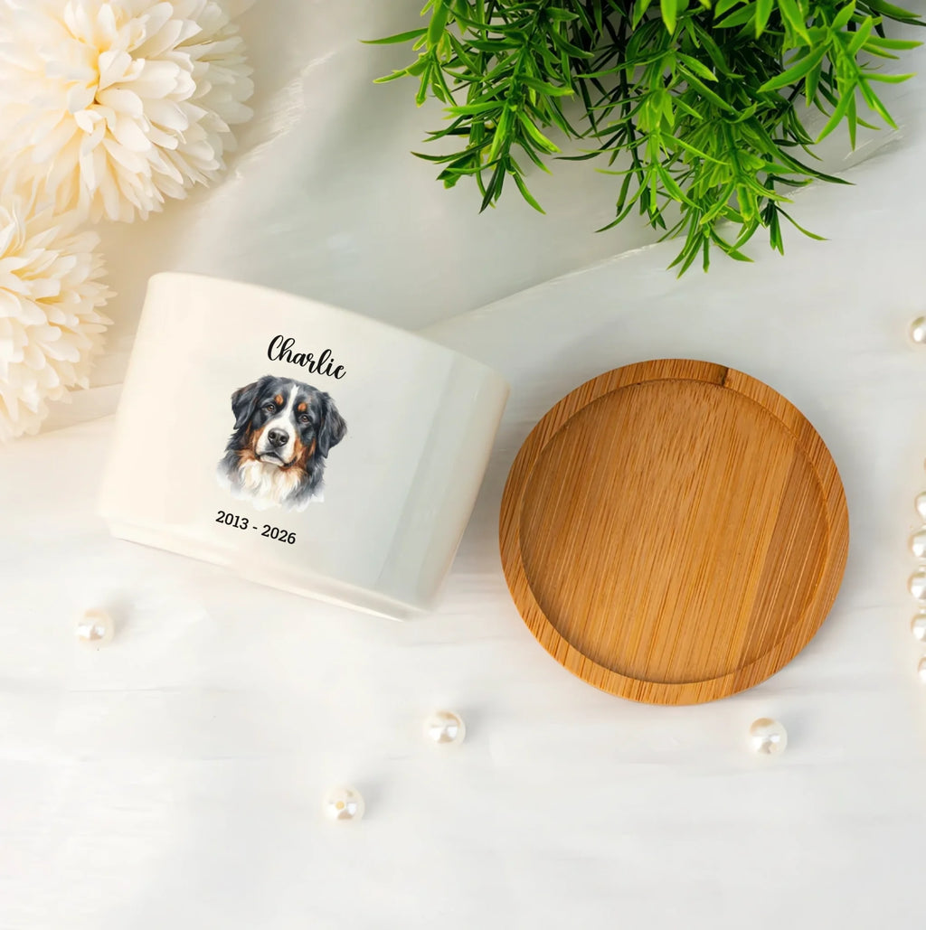 A white ceramic keepsake jar with a bamboo lid, featuring a custom printed dog portrait and the name 'Charlie' on the front. The jar is styled in a flat-lay arrangement with white flowers, green foliage, and small pearls on a white background.