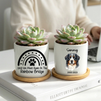 Two white ceramic memorial planters with wooden coasters sit on a white book in a home setting. The left planter has a black rainbow and paw print design with the text 'Until We Meet Again At The Rainbow Bridge'.