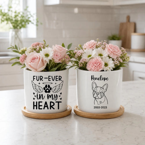 Two white ceramic pet memorial planters with wooden saucers on a kitchen counter. The left planter has black text reading 'FUR-EVER in my HEART' with a paw print and wings, and the right planter has a black line drawing of a French bulldog with the name 'Penelope' and '2003-2023'.