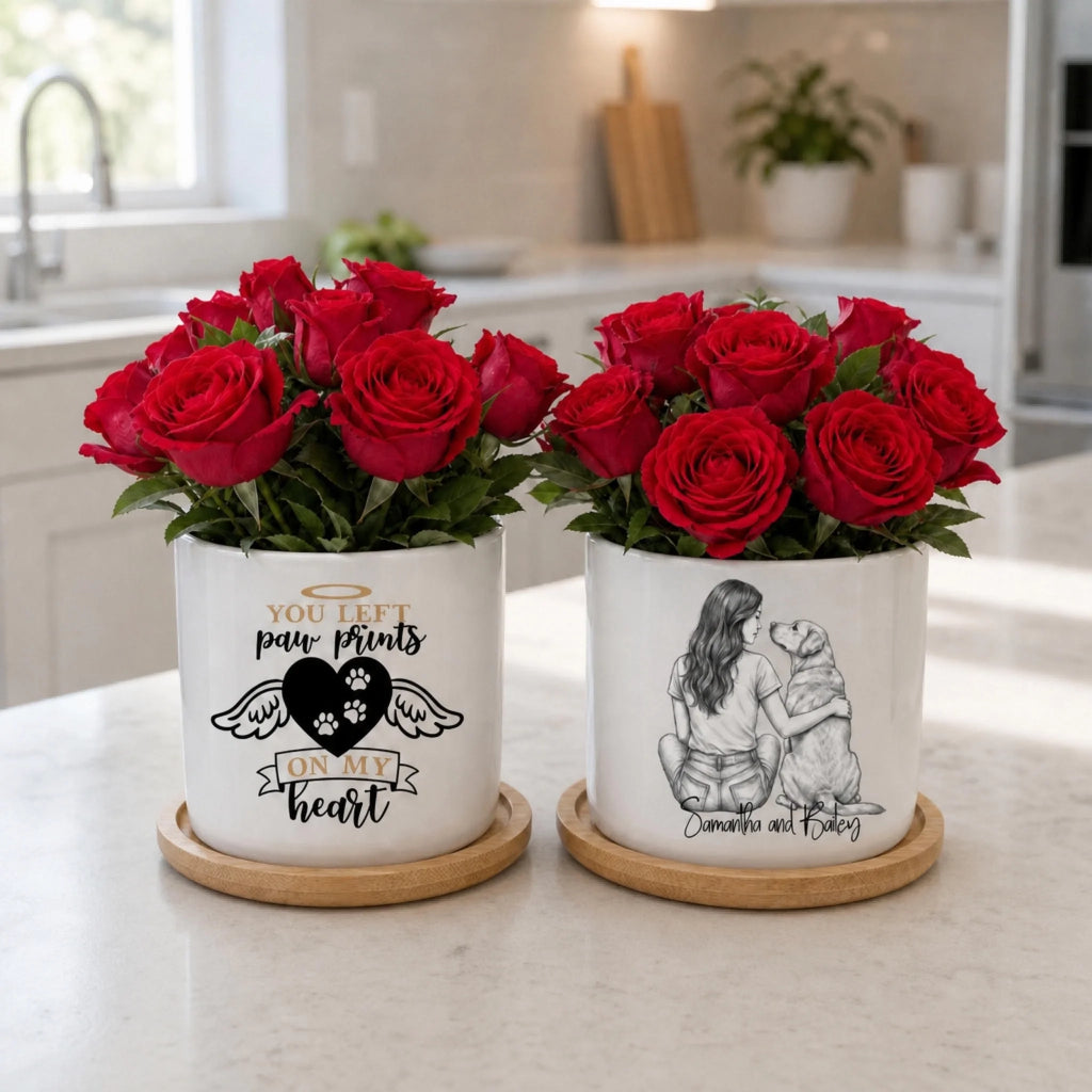 Two white ceramic flower pots with red roses sit on a light marble kitchen countertop. The left pot has a black paw print design with the text 'YOU LEFT paw prints ON ME heart', and the right pot has a black sketch of a woman hugging a dog with the text 'Dedication and bonding'.