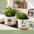 Two white ceramic pet memorial planters with wooden bases sit on a kitchen counter. Each planter contains green herbs and features a black printed design: one says 'You left paw prints and heart' with a winged heart, and the other shows a sketch of a woman holding a dog with the text 'You left paw prints and heart.