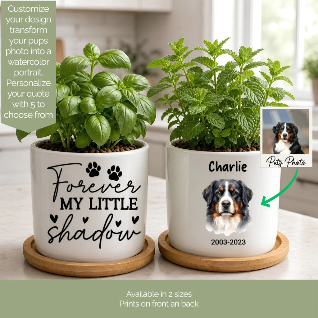 Two white ceramic pet memorial planters with wooden bases, filled with green plants, shown on a kitchen counter. One planter has black script text reading 'Forever My Little Shadow' with paw prints, and the other has a custom dog portrait labeled 'Charlie 2003-2023' with an arrow pointing to a sample pet photo inset.