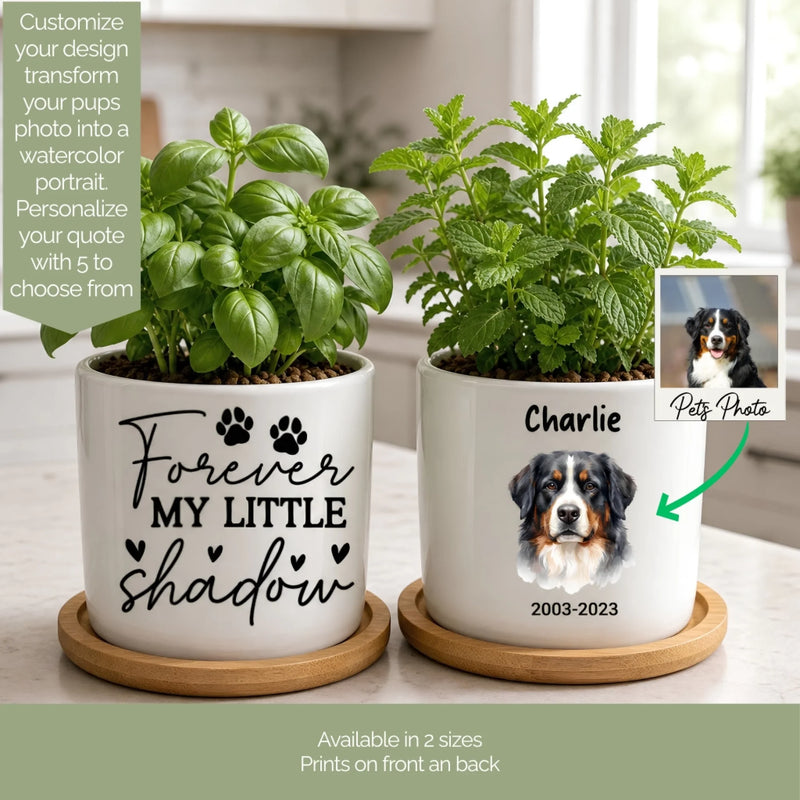 Two white ceramic pet memorial planters with wooden bases, filled with green plants, shown on a kitchen counter. One planter has black script text reading 'Forever My Little Shadow' with paw prints, and the other has a custom dog portrait labeled 'Charlie 2003-2023' with an arrow pointing to a sample pet photo inset.