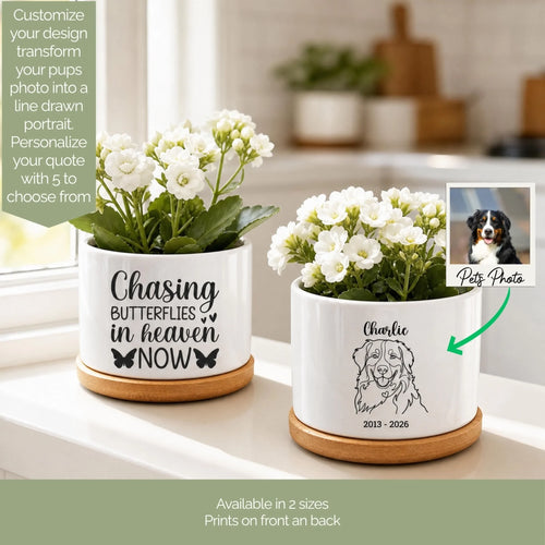 Two white ceramic pet memorial planters with wooden bases displayed on a windowsill. One planter features a customizable quote 'Chasing Butterflies in Heaven Now' with butterfly graphics, and the other is personalized with the name 'Charlie' and a dog illustration.