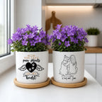 Two white ceramic planters with purple flowers sit on wooden coasters on a white countertop. The left planter has a black heart with paw prints and the text 'YOU LEFT paw prints ON MY heart', and the right planter has a line drawing of a person hugging a dog with the text 'Rose and Jane'.