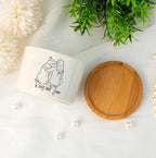 A white ceramic candle jar with a black line art illustration of two hugging figures and the text 'Kong and Jane' on the front, shown next to its round wooden lid on a white surface with decorative flowers, greenery, and pearls.