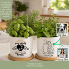 Two white ceramic pet memorial planters filled with green herbs, shown on wooden coasters in a bright kitchen setting. The left planter has a customizable black graphic with wings, a heart, paw prints, and the text 'YOU LEFT paw prints ON MY heart'.