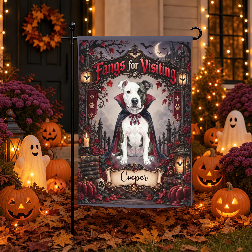 A Halloween-themed garden flag featuring a white pitbull-type dog dressed as a vampire with the text 'Fangs for Visiting' and the name 'Cooper' at the bottom, displayed on a black flagpole in a decorated front yard with pumpkins, ghosts, and warm string lights.